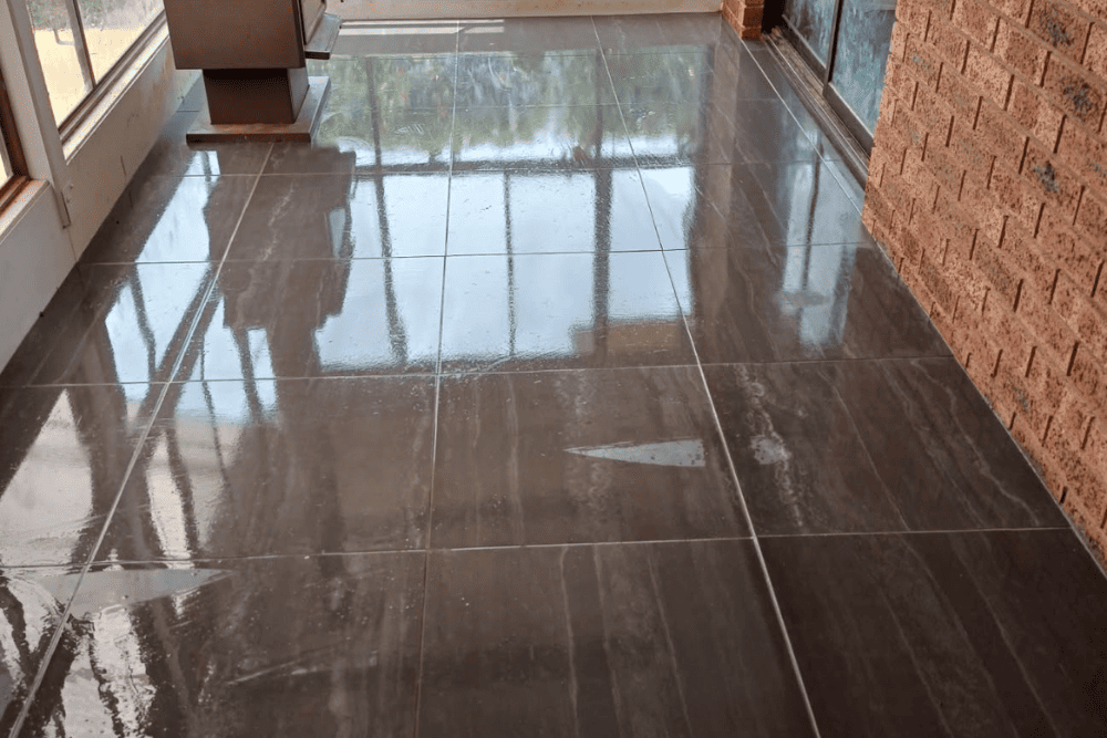 Clean, highly polished dark floor tiles indoors reflecting sunlight near a brick wall-